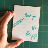 white greeting card with teal illustrated paper lines, pencil shavings, and a pencil next to hand lettering that reads "thank you" photographed being hand-held