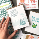 white greeting card with a blue and teal illustration of an a-frame cabin and tree line of pines. hand lettering above the house reads "sending love + light this holiday season" shown as a boxed set hand-held amongst other boxed holiday cards