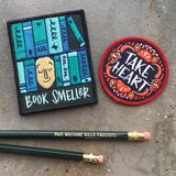 Two patches, one a blue book smeller and one a red and black patterned take heart above two green pencils that say, "this machine kills fascists."