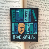 A rectangle patch with a face surrounded by various shades of blue books with text under it that says, "book smeller" on top the text from a book.
