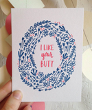 Greeting card and kraft paper envelope. Illustrated wreath of navy flowers surround red/pink text that reads, "I like your butt."