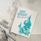 Greeting card and kraft paper envelope. Reads, "Happy birthday, sorry it took so long." Illustration of snail riding a sloth (who is eating cake) riding a tortoise all in festive birthday hats. Drawing in shades of teal.