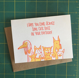 Greeting card and kraft envelope. Hand-written text at top reads, "I hope you come across some cute dogs on your birthday." Illustration below of a group of dogs of various breeds and sizes. Colors are red and orange.