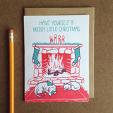 A greeting card and envelope featuring an illustration in green and red of a fire place with stockings hanging and a cat and dog laying in front of it. The text abouve reads "have yourself a merry little Christmas".