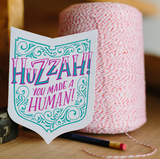 A shield shape die-cut greeting card with kraft paper envelope. Festive hand-drawn text that reads "huzzah! you made a human!" in purple and teal, surrounded by teal flourishes.