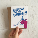 greeting card and kraft paper envelope. Hand-written text in blue reads, "Holy cow! It's your birthday?!" above illustration of cow in party hat with noise maker.