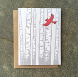 A greeting card with birch trees and a cardinal flying in front of it.