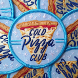 a pile of blue patches that say, "cold pizza club" against a slice of pizza.