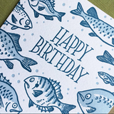 Greeting card with kraft envelope. Reads "happy birthday" in hand-written, serif font, surrounded by illustrated fish of various sizes and shapes. Shades of blue.