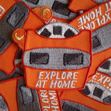A pile of embroidered iron on patches that are die cut in the shape of a vintage orange view finder with text that says "Explore At Home."