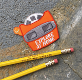 An embroidered iron on patch die cut in the shape of a vintage orange view finder with text that says "Explore At Home" besides a yellow pencil reading, "Oh baby you you got what I nee eed"