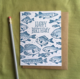 Greeting card with kraft envelope. Reads "happy birthday" in hand-written, serif font, surrounded by illustrated fish of various sizes and shapes. Shades of blue.