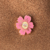 An enamel pin with a butterfly back in the shape of a pink flower with a very silly indifferent face in the center on top of corkboard