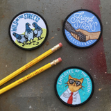 Three iron on embroidered patches, from left to right: a patch with pigeons on it that says "wild in the streets", a patch with a dog paw and hand embracing with text that says "friend to strays", and a cat named bill in a button up shirt and glasses with text above reading "employee of the month"