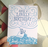 Greeting card and kraft paper envelope. Illustration of rat on top of garbage pile happily saying "have a lovely birthday" in hand-written delicate font surrounded by swirls. Drawn in shades of blue.