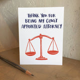 white greeting card that reads "thank you for being my court appointed attorney" in navy above a red illustration of old school scales photographed with a pencil