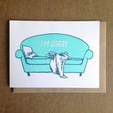 white greeting card featuring an illustrated teal couch with a pillow and blanket slumped on it. white text on the couch reads "I'm sorry"