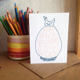 white greeting card featuring a blue illustration of a hen sitting atop an extremely large egg. orange hand-lettering on the egg reads "mom - how do you make it look so easy?" pictured with a colorful cup of pencils