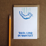 white greeting card featuring an illustrated falling cat which reads "you'll land on your feet" shown next to a pencil