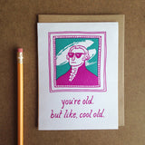 greeting card with a pink and teal illustration of Gilbert Stuart which reads "you're old, but like, cool old" shown next to a pencil