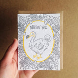 Greeeting card and kraft paper envelope. Grey, illustrated tropical leaves surrounding dodo bird with text above him that reads, "Missin' you."