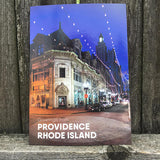 postcard photograph of iconic downtown Westminster Street lit up at night with white text on the bottom that reads "Providence, Rhod Island" photographed in front of a fence