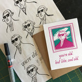 greeting card with a pink and teal illustration of Gilbert Stuart which reads "you're old, but like, cool old" shown with design sketches and a pen
