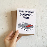 white greeting card with an illustrated double bed benath navy text that reads "two words: conjugal visit" photographed hand-held in fron of a wall