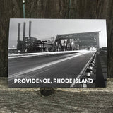black and white postcard photograph of night-time traffic driving over a downtown providence bridge with the iconic power station smoke stacks visable in the background. white text on the bottom reads "greetings from providence, rhode island"