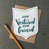 badge-shaped greeting card with a light blue illustration of a mountain range behind green hand-lettering that reads "nothing ventured nothing gained" photographed with a pencil