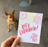 Greeting card and kraft paper envelope. Text reads, "Let's celebrate!" Illustration of fireworks with dog below, shaking, holding a sign that says, "quietly." Photographed hand-held with a dog viasble in the background