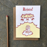 white greeting card featuring a maroon and yellow illustration of a carton of eggnog ascending twards trumpets in the clouds. hand lettering at the top reads "rejoice!" photographed with a pencil