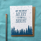 white greeting card with a navy pine tree design beneath hand-lettering that reads "may your days be merry and bright" amongst a pale blue starry sky photographed with a pencil