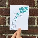 Greeting card and craft envelope that reads "welcome peanut!" in teal, hand-written type in upper right corner. Illustration of elephant trunk shaking rattle. Photographed hand-held in front of a brick wall