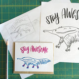 white greeting card with an illustrated ant-eater on rollerskates beneath hand-lettering that reads "stay awesome" photographed with a pencil and design sketches