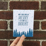 white greeting card with a navy pine tree design beneath hand-lettering that reads "may your days be merry and bright" amongst a pale blue starry sky photographed hand-held in front of a brick wall