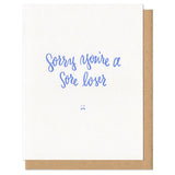 white greeting card with blue text that reads "sorry you're a sore loser" above a small frowny face drawing