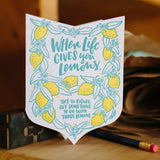 Die cut badge-shaped greeting card that reads "when life give you lemons, try to figure out something to do with those lemons" in teal text surrounded by illuatrated lemons and leaves photographed amongst studio equipment