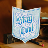 badge-shaped greeting card printed in blue and teal with a dot pattern surrounding handlettering that reads "stay cool" photographed with a pencil and letterpress blocks