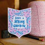 Die-cut, shield shaped greeting card. Handwritten type in navy reads, "Still a spring chicken." Surrounded by navy and purple flowers and leaves, photographed with a pencil and spool of string