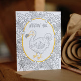 Greeeting card and kraft paper envelope. Grey, illustrated tropical leaves surrounding dodo bird with text above him that reads, "Missin' you."