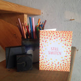 greeting card with a red and yellow star patter surrounded hand-letterinf that reads "still smitten" photographed with a cup of pencils and letterpress type