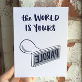 white greeting card that reads "the world is yours" in navy text above an illustration of a "parole" stamp photographed being hand-held infront of a brick wall