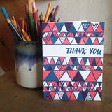white greeting card with a red and blue illustrated triangles pattern and hand lettering that reads "thank you" photographed next to a cup of pencils