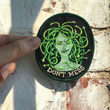 A patch of a green medusa with snakes for hair and "don't mess" below her held against a white brick wall.