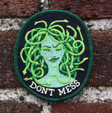 A patch of a green medusa with snakes for hair and "don't mess" below her held against a red brick wall.