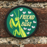 An iron on embroidered circle patch with a black outline and a forest green background with a praying mantis, flies, and text that reads "friend to bugs" on a brick background