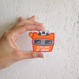 An embroidered iron on patch die cut in the shape of a vintage orange view finder with text that says "Explore At Home" being held against a white wood wall
