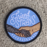 A circle iron on embroidered patch with a black outline and a light blue background with a dog's paw holding a human's hand with scripted text above it reading "friend to strays" on the concrete ground