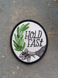 An oval iron on embroidered patch featuring an eagle's claw holding an olive branch with script text next to it that says, "Hold fast."